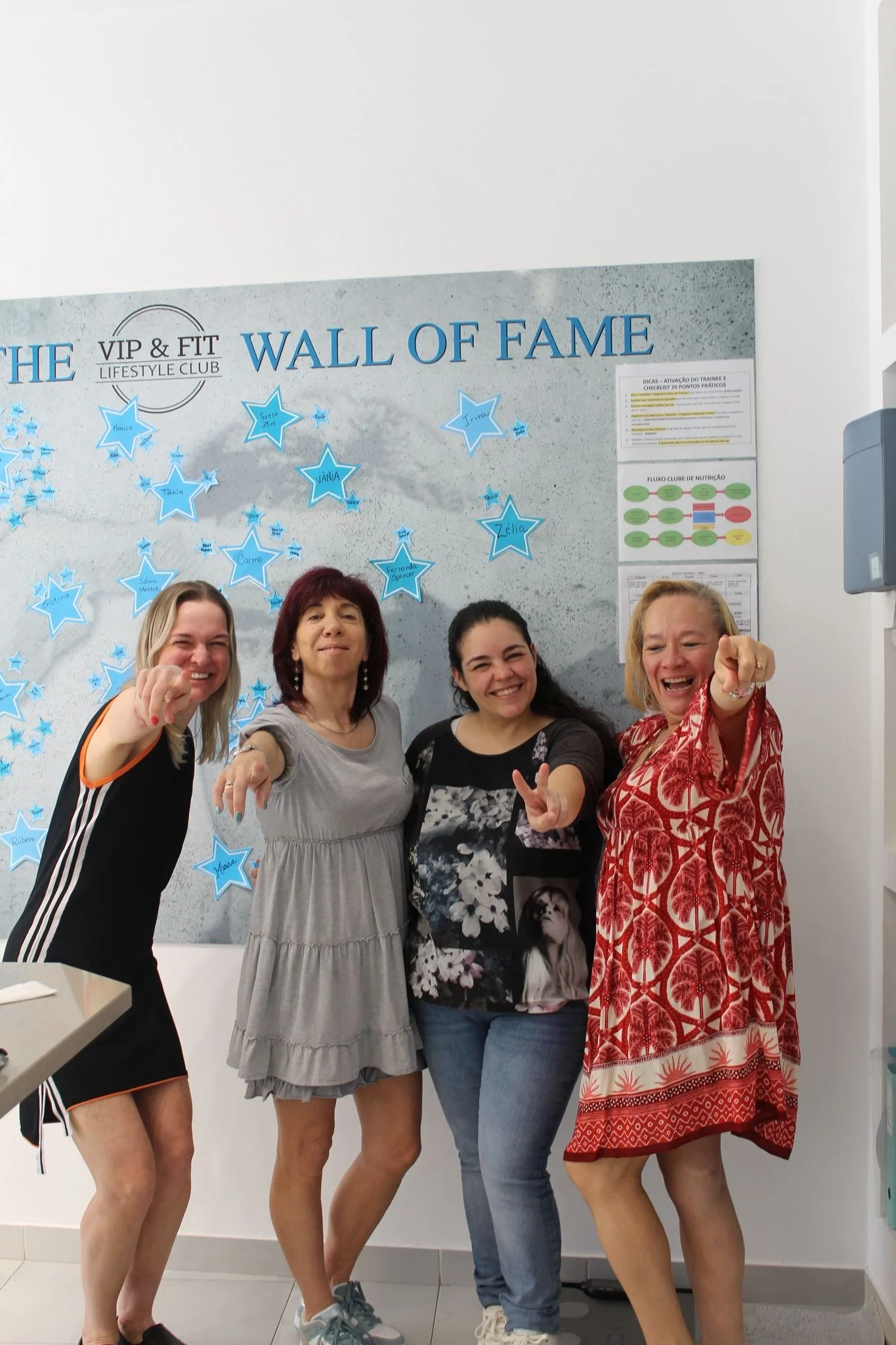 Quatro mulheres sorrindo e apontando para a câmera em frente a um mural intitulado 'Wall of Fame' com estrelas azuis e nomes escritos nelas.