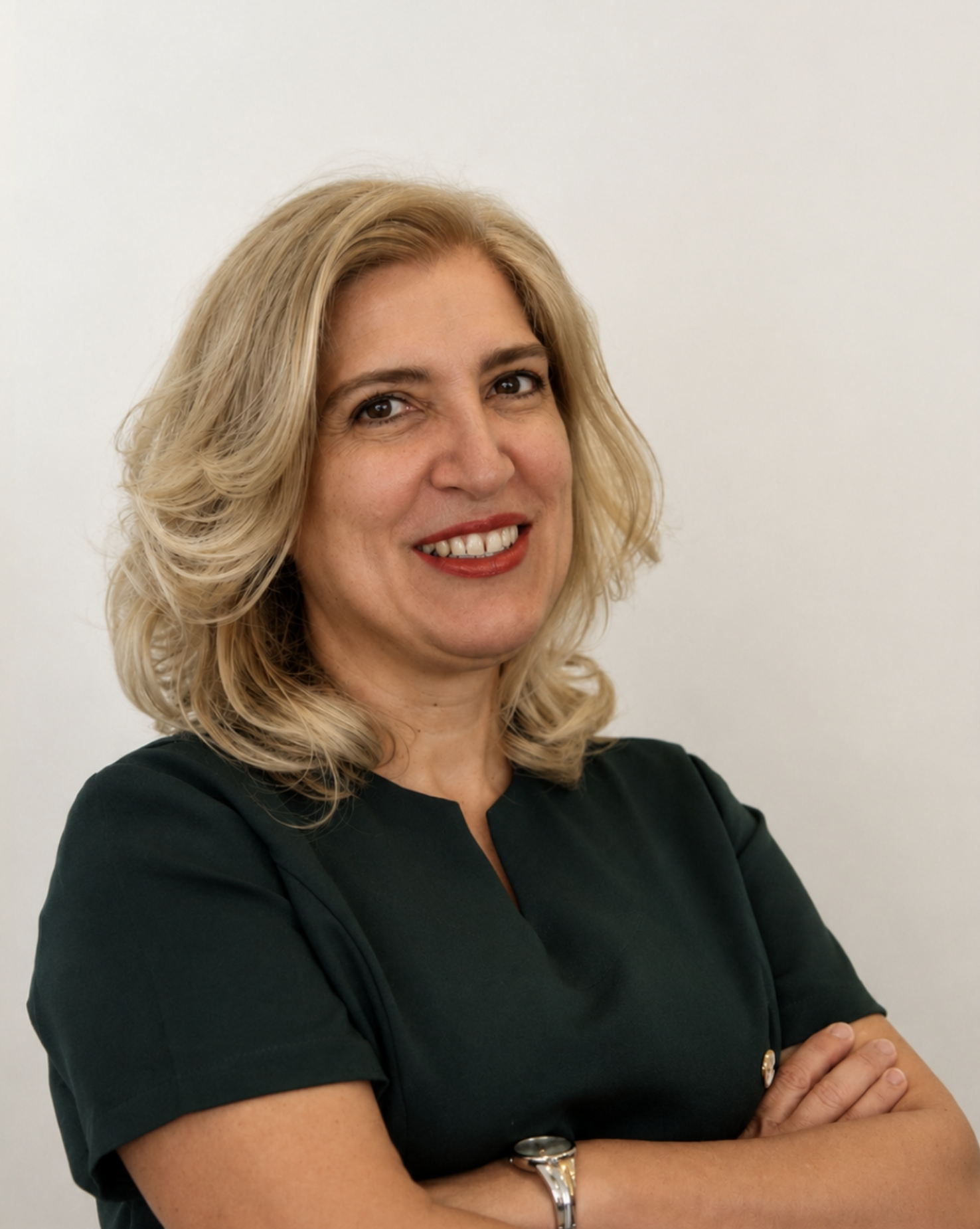 Mulher com cabelo loiro, pele clara, vestindo uma blusa verde escura, com braço cruzado e sorrindo, contra fundo branco.