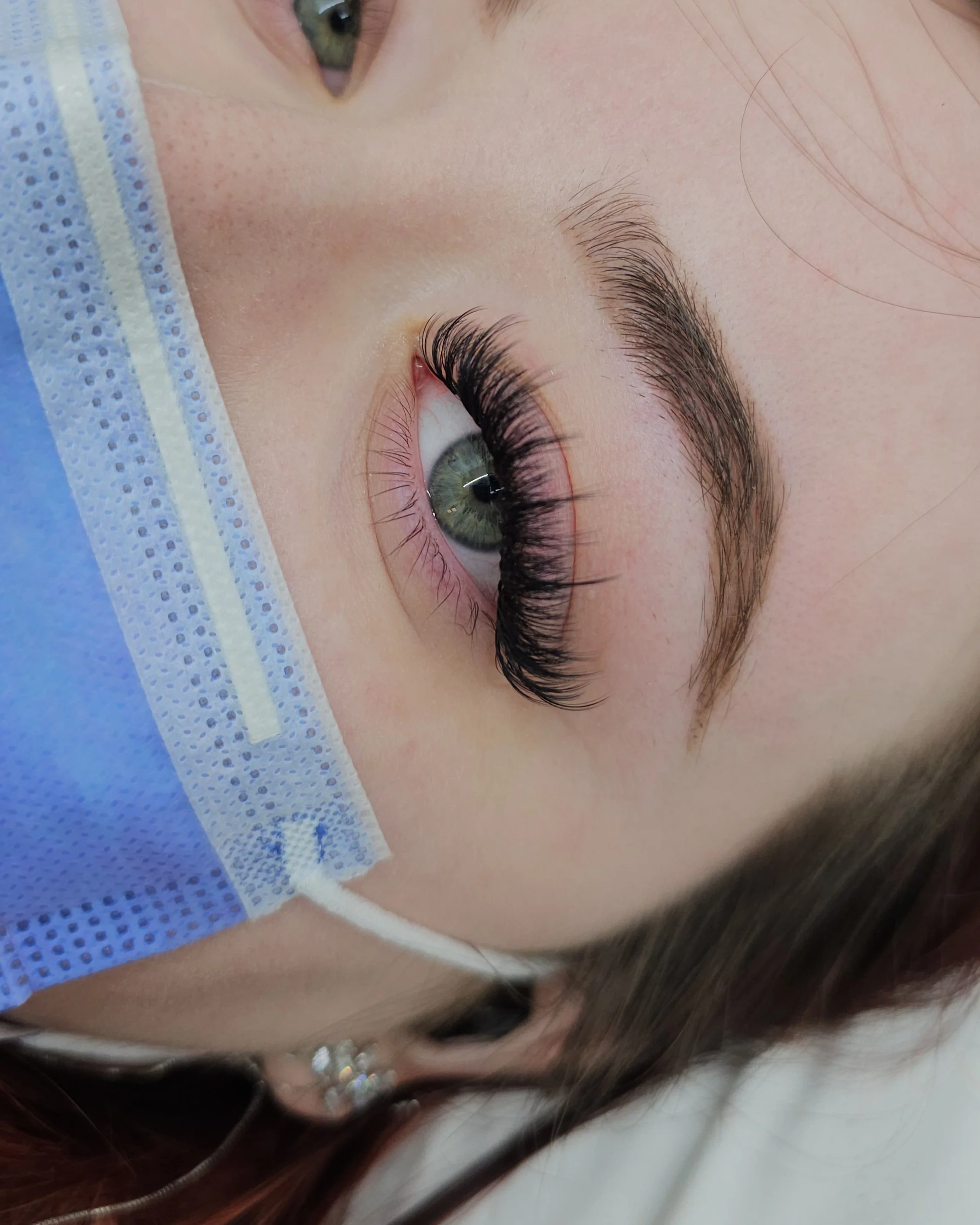 Close-up of a person's eye with freshly applied eyelash extensions, wearing a blue face mask.