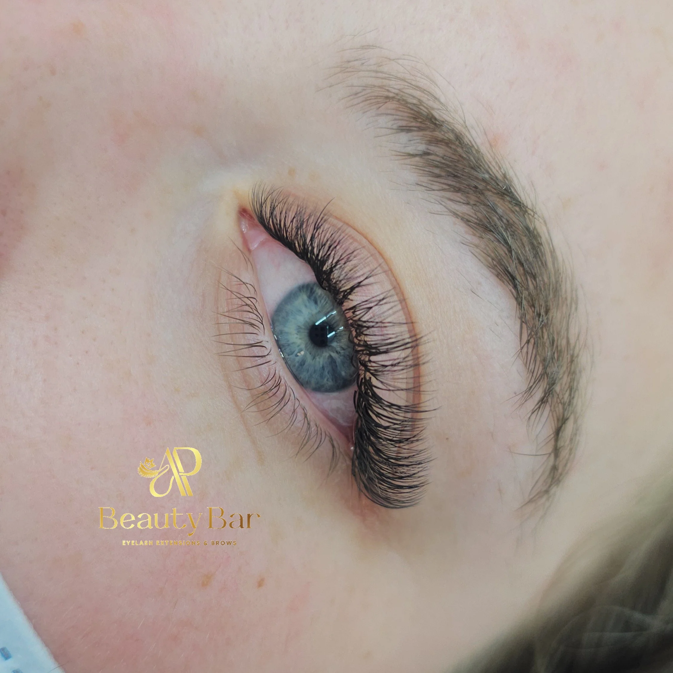Close-up of a woman's blue eye with long, thick eyelashes, and well-groomed eyebrow. The image includes the logo of a beauty bar specializing in eyelash extensions and brows.