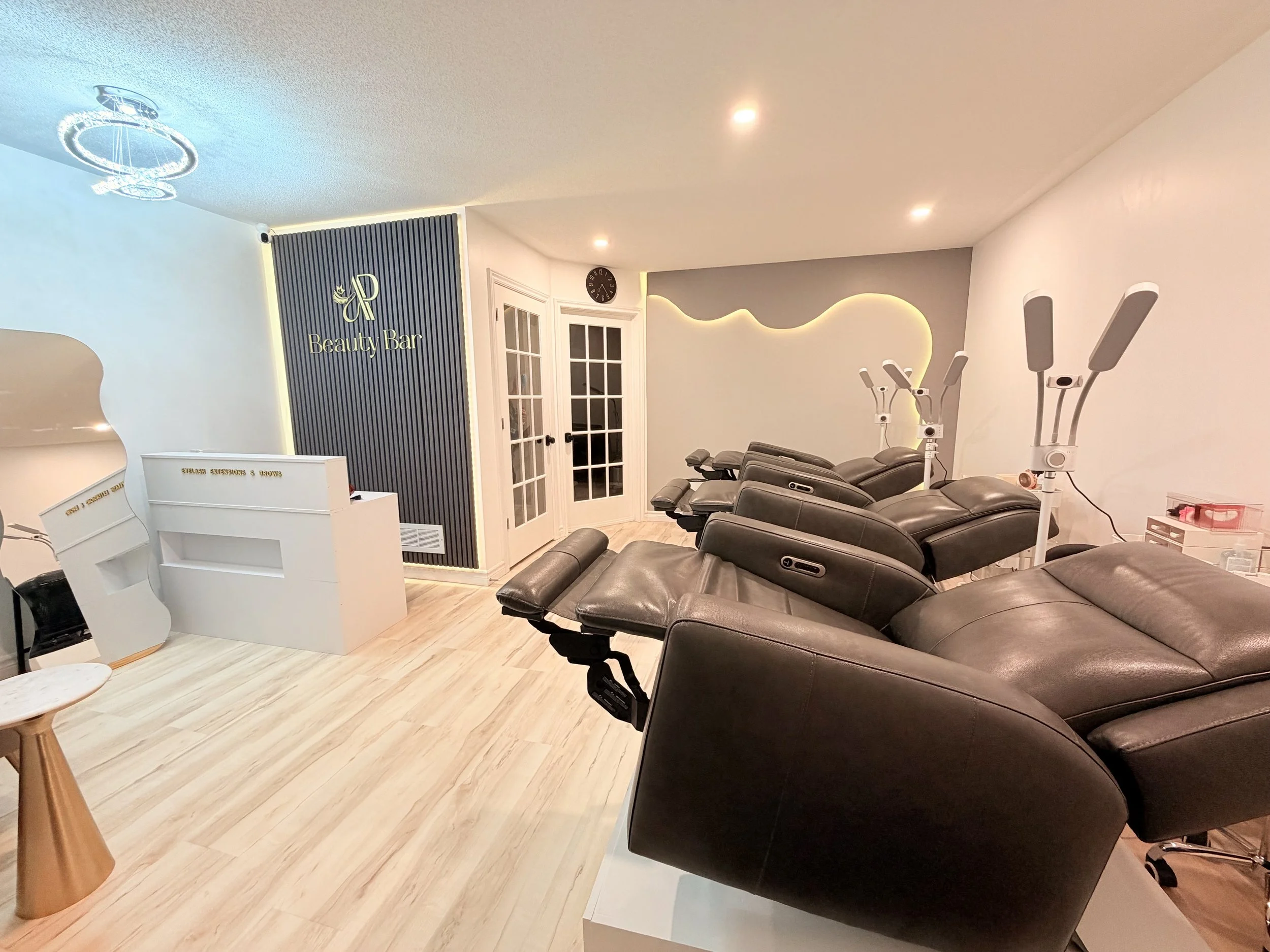 Interior of a beauty salon with four black adjustable chairs, facial and massage beds, and skincare equipment against a white wall with decorative lighting.