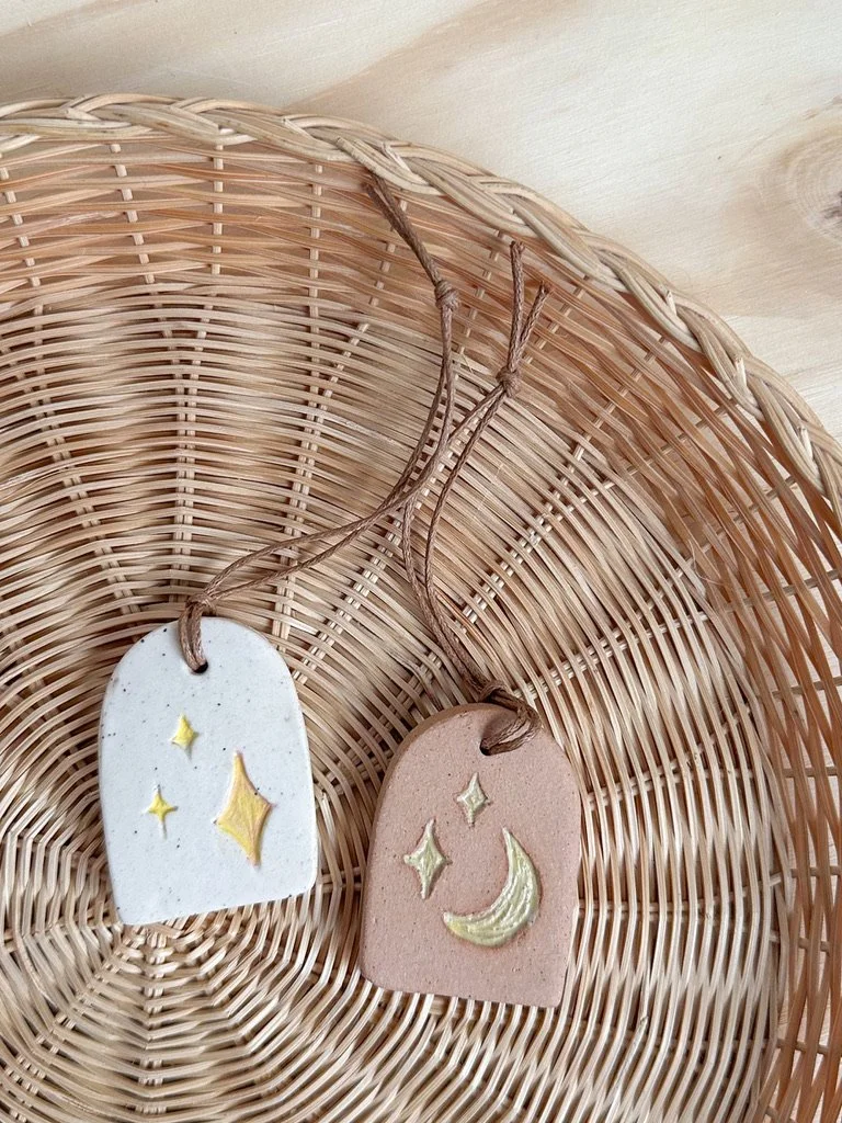 Two decorative pottery tags with celestial designs, one with yellow stars and the other with a yellow crescent moon, hanging on a woven basket.