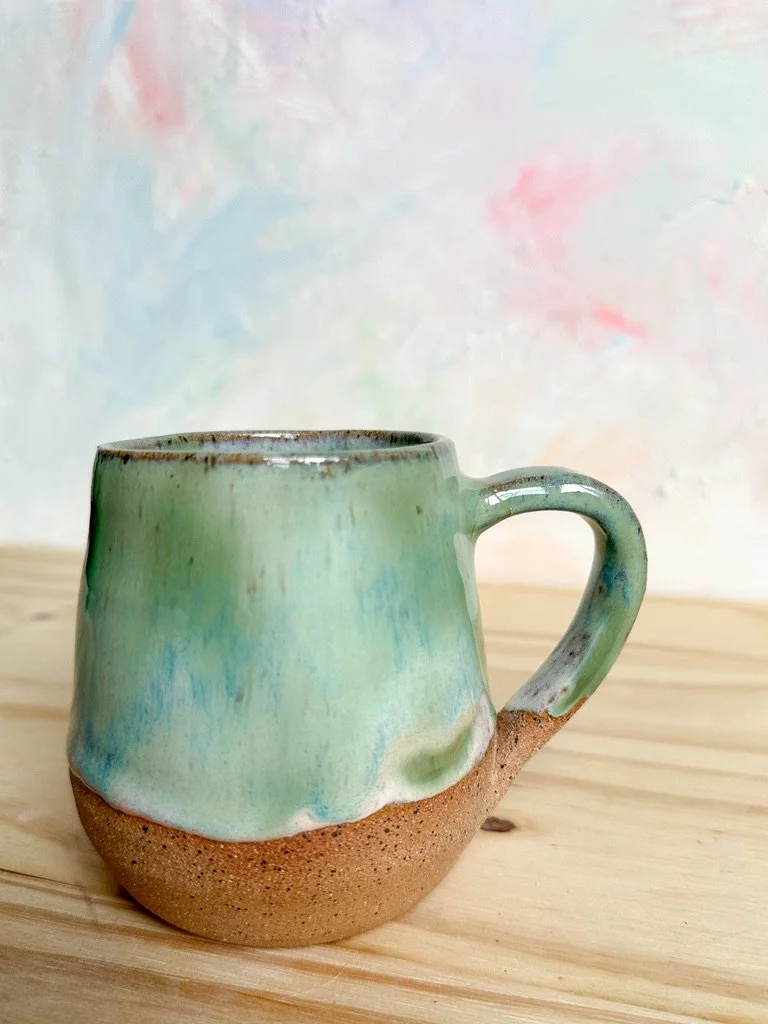 A handmade ceramic mug with a glazed light green and blue top that fades into a natural unglazed clay bottom, sitting on a wooden surface with a pastel abstract background.