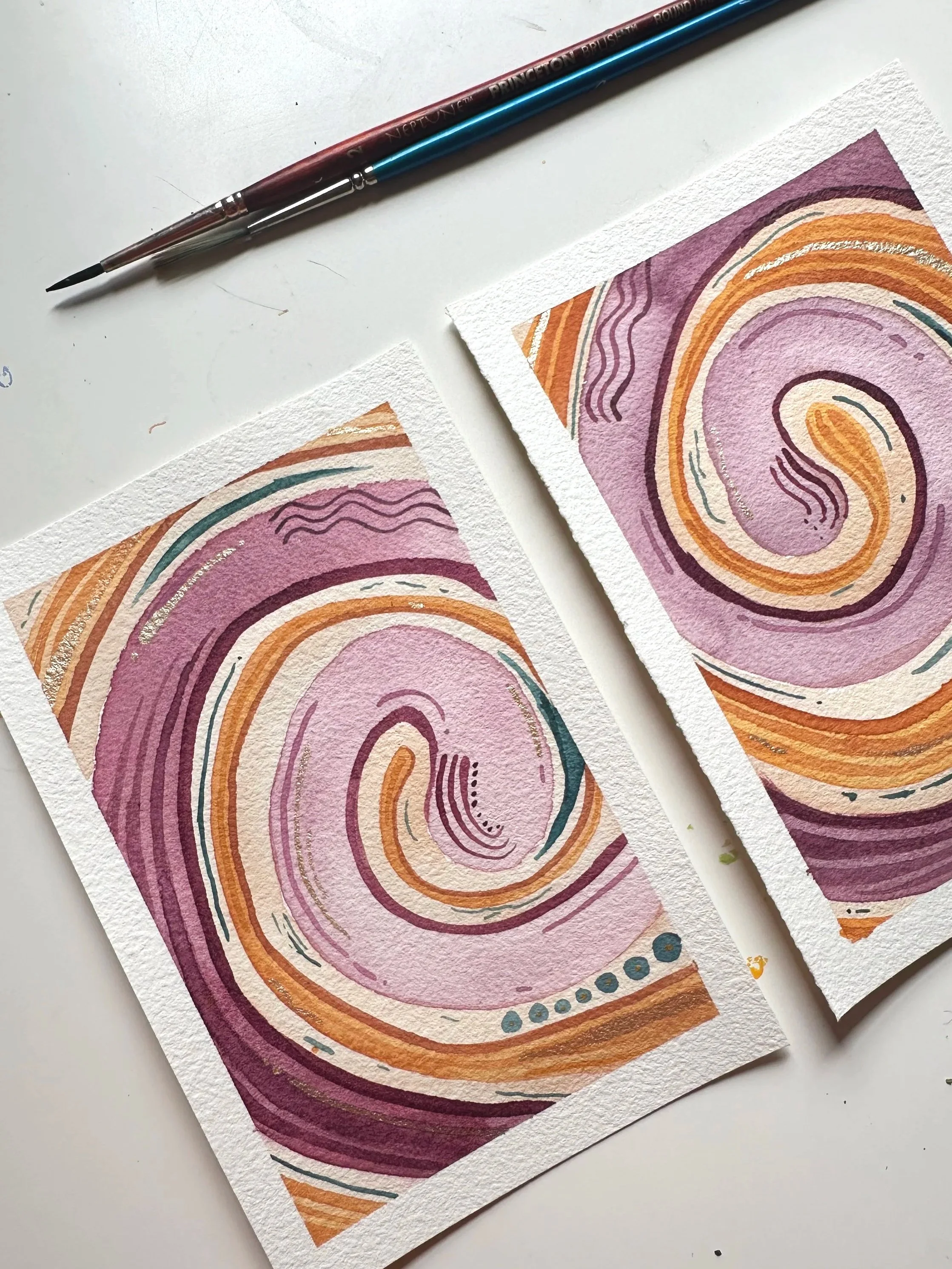 Two small spiral watercolor portraits sit on a white desk with paintbrushes. The art is pinks and oranges.