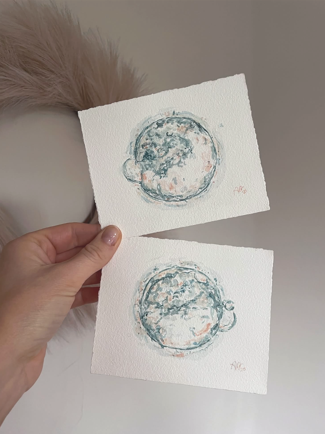 A hand holds two small watercolor pantings of embryo images in blue and peach.