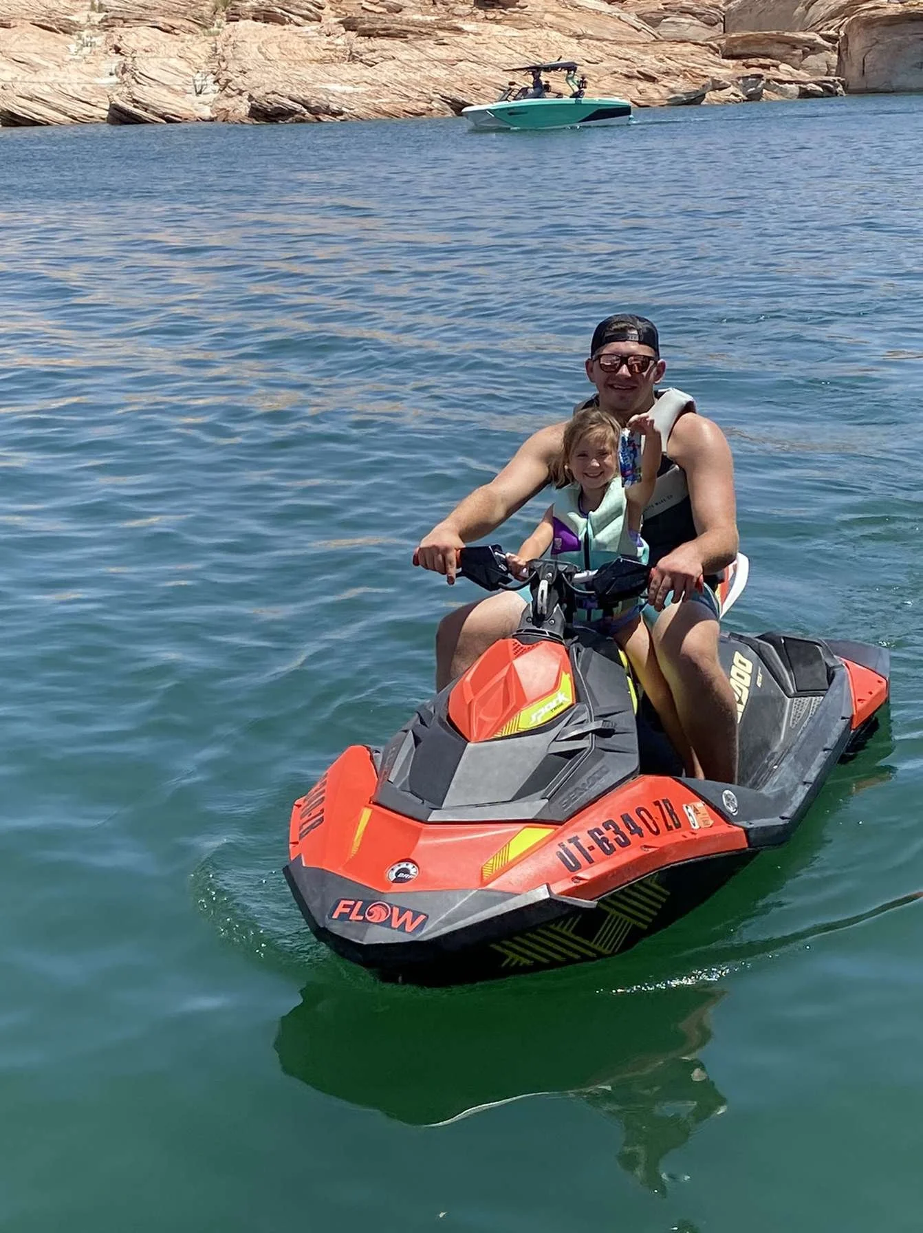 A man and a young girl riding a red and black jet ski on calm water, wearing life jackets, with rocky terrain in the background.