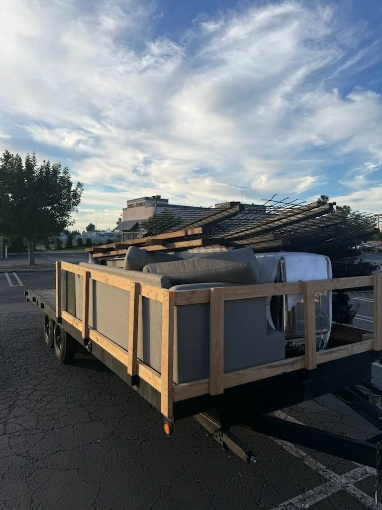 A trailer loaded with various building materials, including metal frames, pipes, and wooden planks, parked in a lot with a partly cloudy sky overhead.