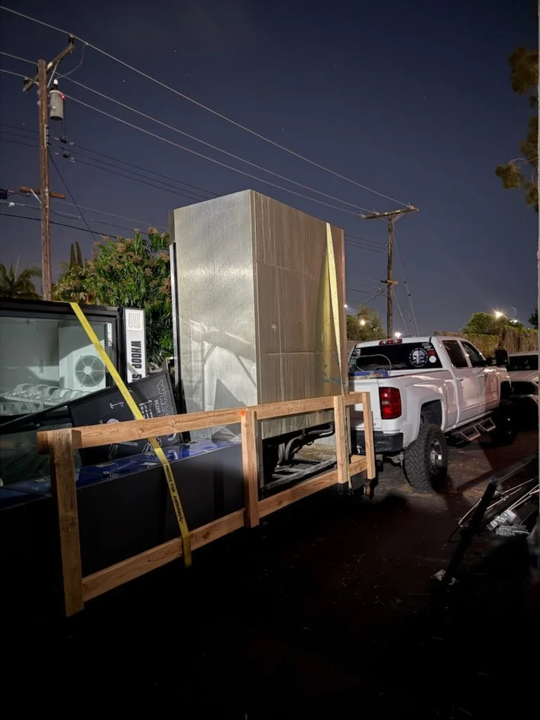 A white pickup truck parked beside a trailer carrying a large metallic cabinet or refrigerator, with some disassembled electronic equipment and a wooden frame, at night.