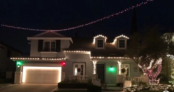 A house decorated with white holiday lights outlining the roof, windows, and porch. There are green and red lights on the garage, and a string of multicolored lights overhead. A pink flowering tree is lit up on the right side.