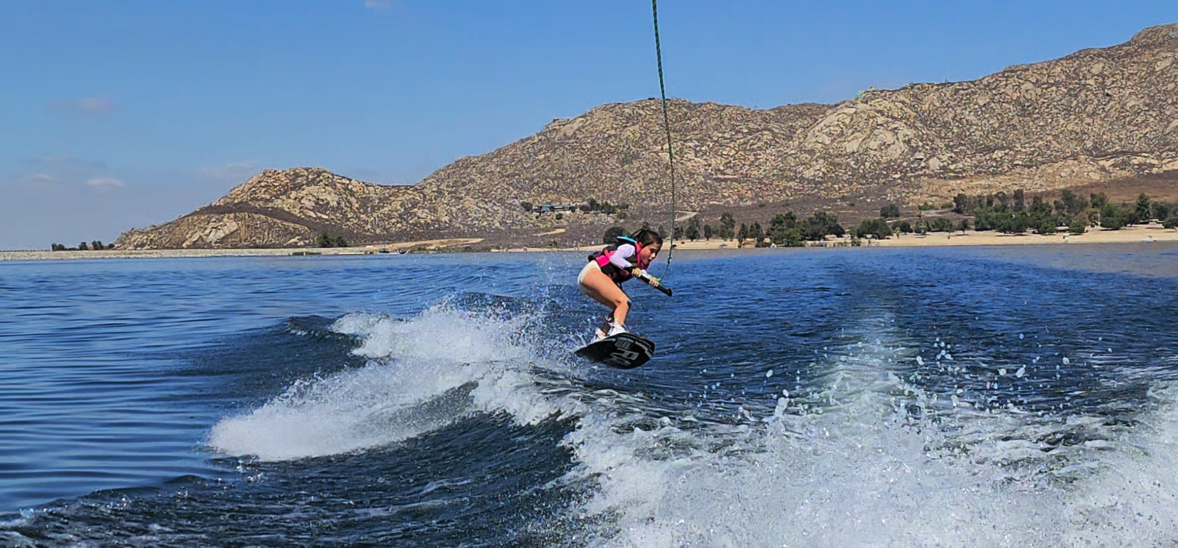 A young girl wakeboarding on a lake with rocky mountains and a beach in the background, wearing a life jacket and holding a handle connected to a cable.