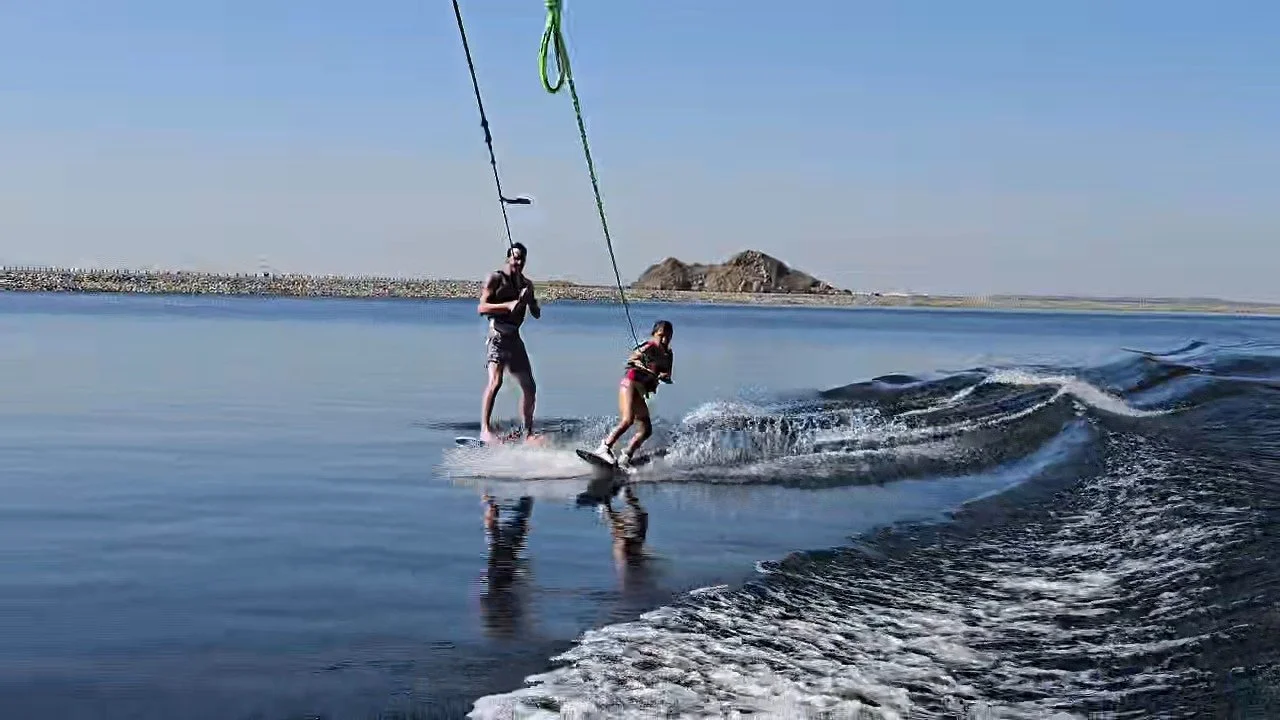 Two people wakeboarding on a calm body of water with a clear blue sky and distant landforms in the background.