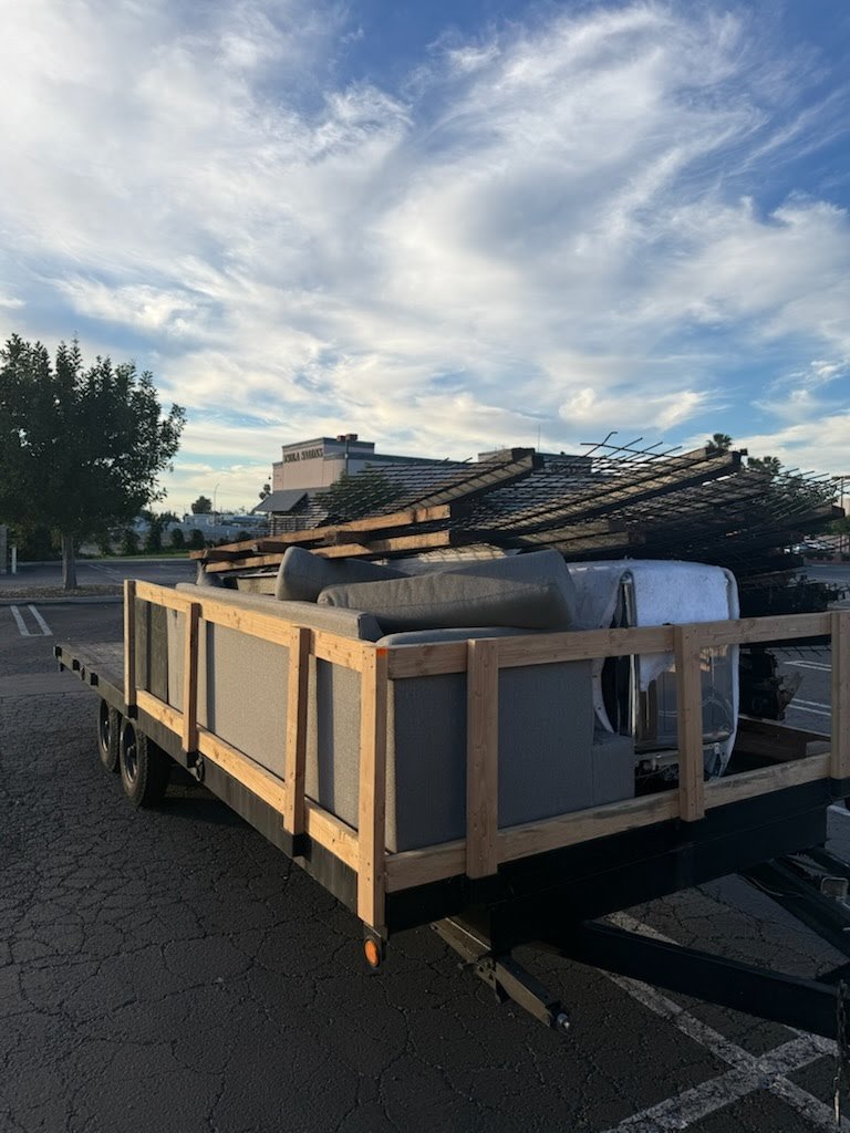 Flatbed trailer parked in a lot with a load of building materials, including wooden planks, metal rods, and insulation rolls, under a partly cloudy sky.