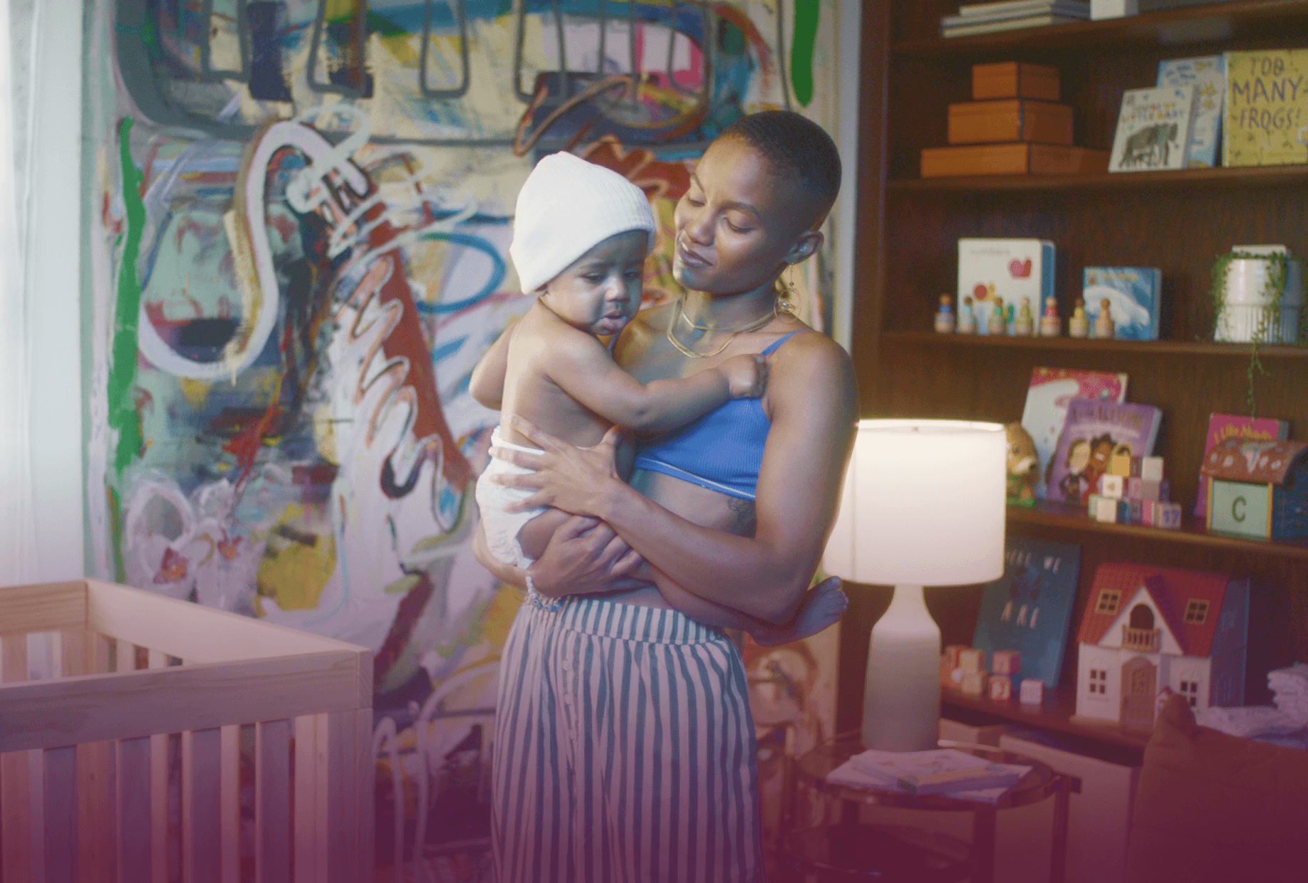 A woman holding a young child in a room with colorful abstract art on the wall, bookshelves with children's books and toys, and a white lamp on a small table.