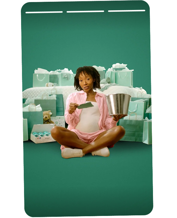 A woman sitting cross-legged on the floor surrounded by boxes of tissues in a room decorated with tissues and related items, holding a piece of tissue in one hand and an empty metal bucket in the other, with a distressed or overwhelmed expression.