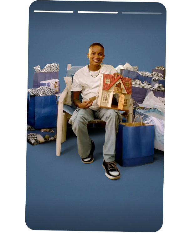 Person sitting on a bench holding a toy dollhouse, surrounded by shopping bags and gifts, in front of a blue background.