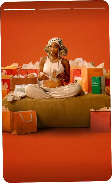 A woman sitting cross-legged on a couch surrounded by shopping bags, holding a gift card, with a warm orange background.