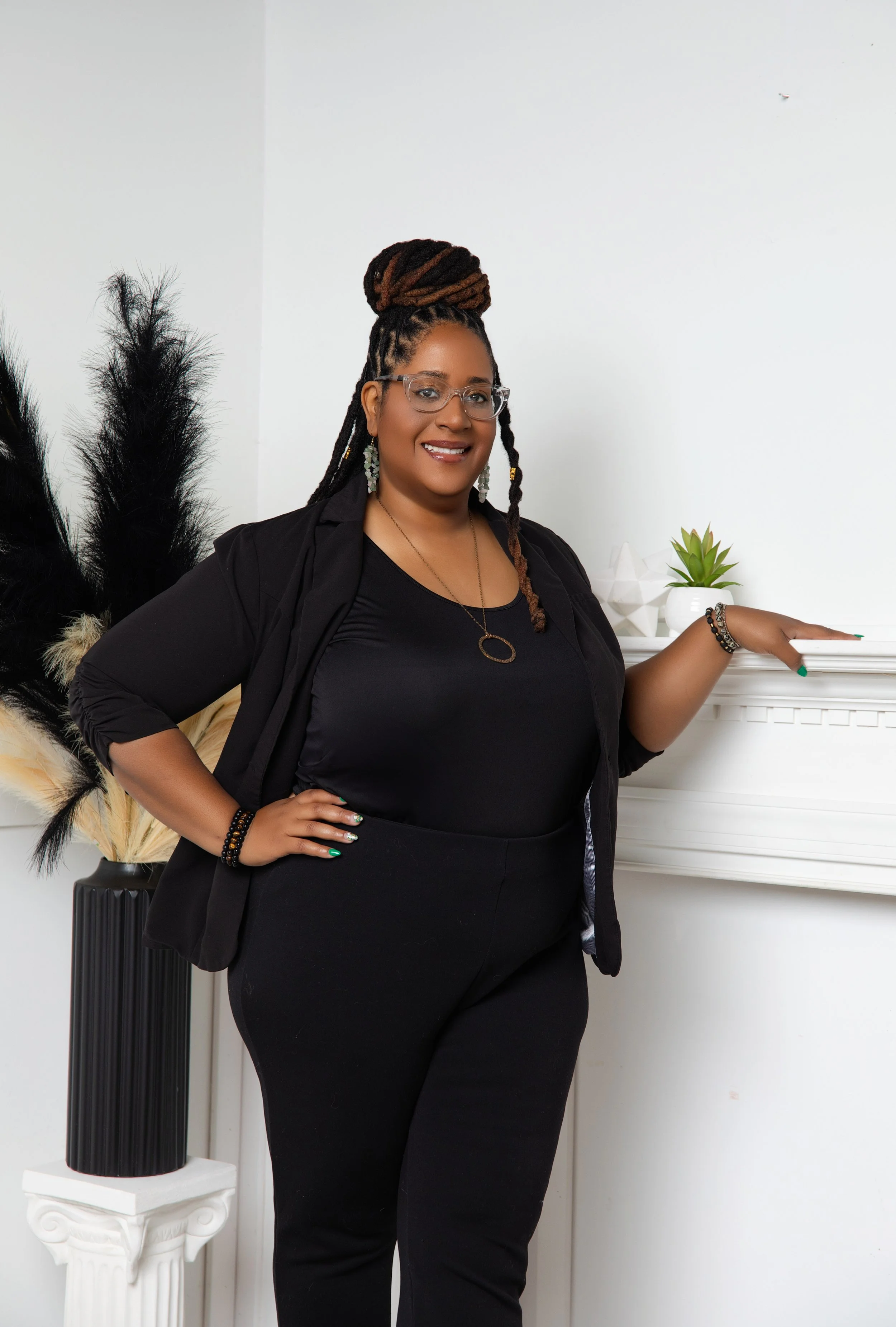A woman with glasses and long dreadlocks styled in a bun, wearing a black blazer over a black top and black pants, standing indoors near a white fireplace mantel with a small potted succulent plant and decorative objects, smiling at the camera.