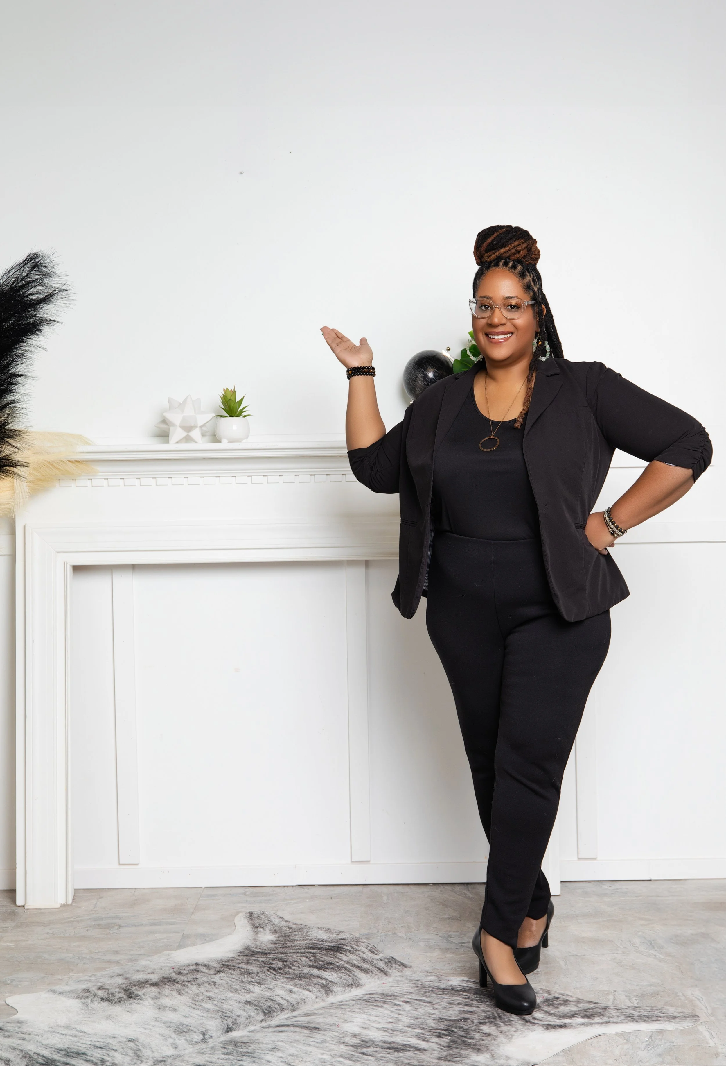 A woman in a black suit standing in front of a white wall, smiling, with one arm raised and the other on her waist, surrounded by decorative objects on a white mantle.