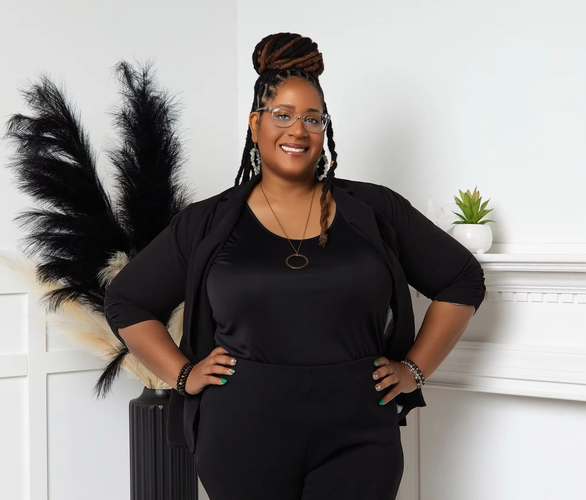 A woman with glasses, styled braids, and dreadlocks in a bun, dressed in black, stands confidently in front of a white wall with a decorative black feather arrangement and a potted plant on a white mantel.