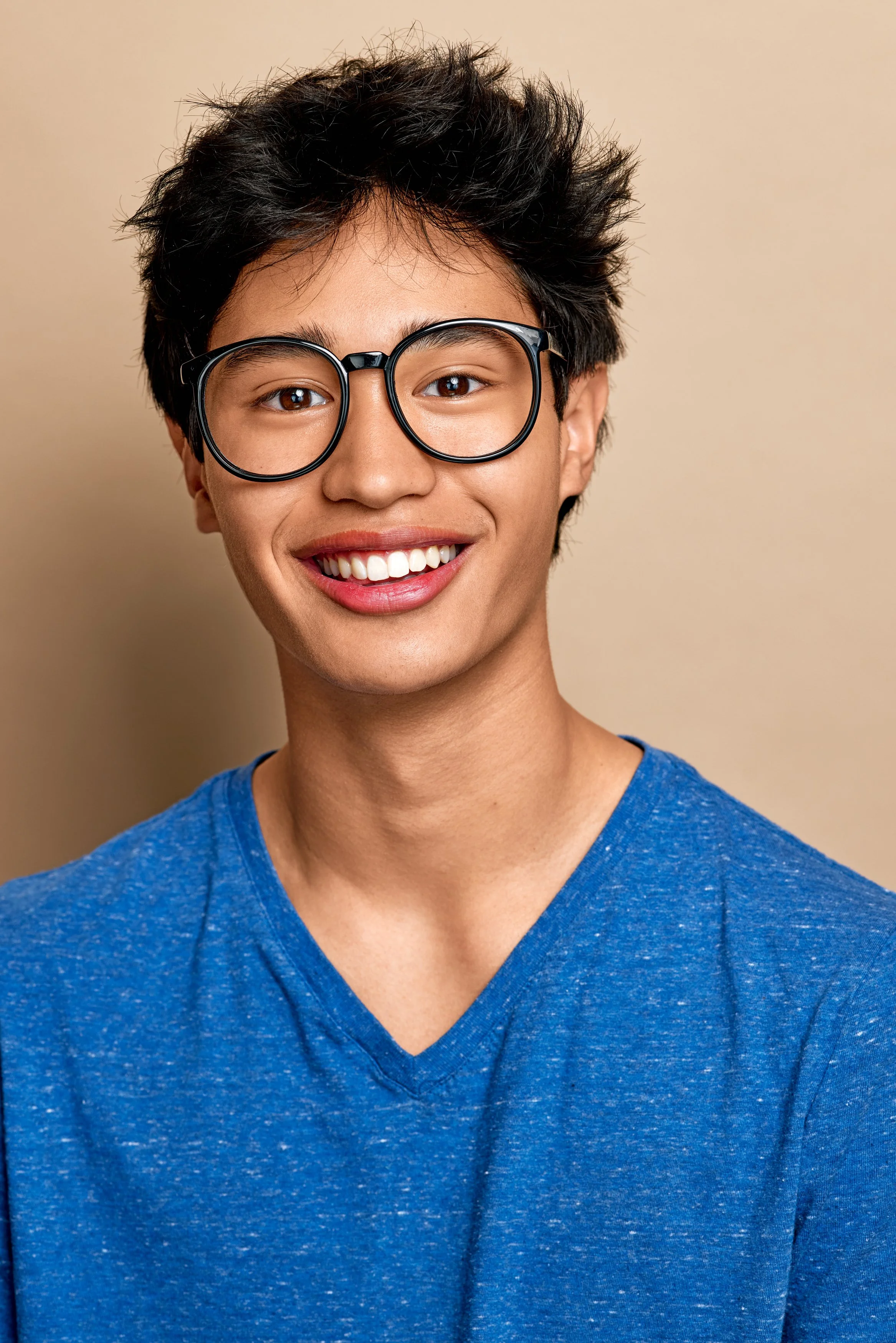 Close-up of a young man with dark, curly hair, wearing round eyeglasses and a blue V-neck shirt, smiling against a beige background.