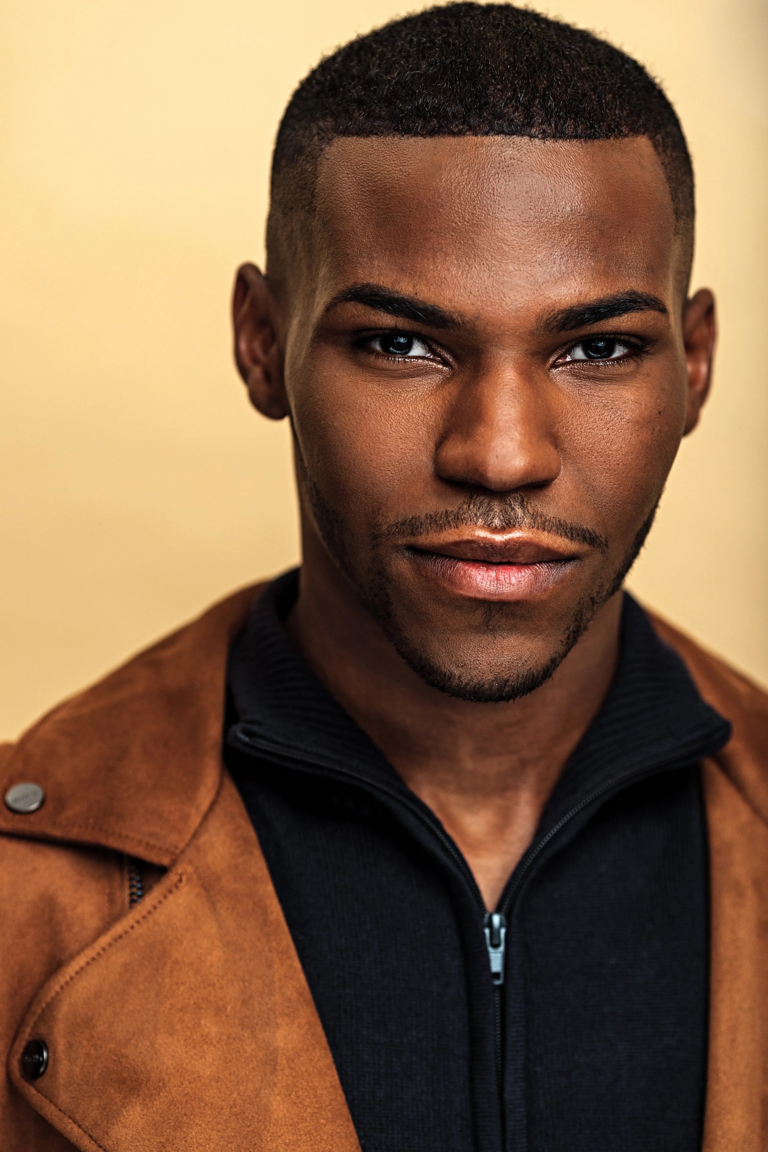 Close-up portrait of a young man with short curly hair, brown skin, piercing blue eyes, wearing a black zip-up jacket and a brown coat over his shoulders, against a beige background.