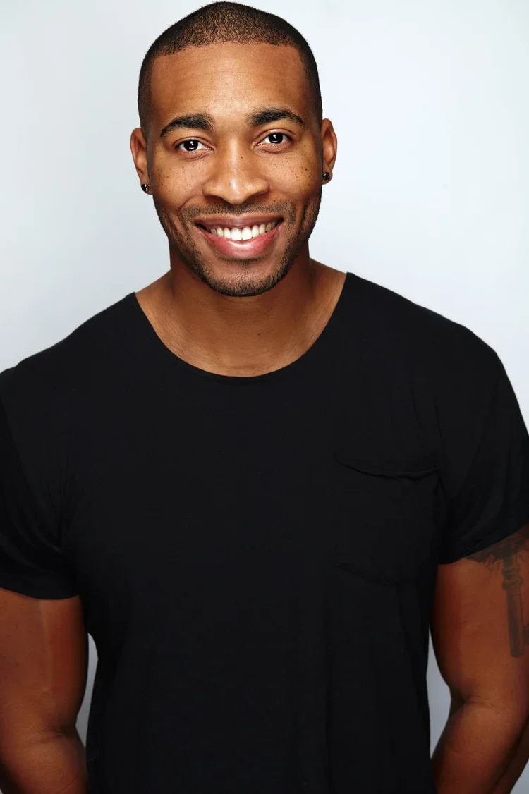 A smiling Black man with short hair, wearing a black t-shirt, standing against a light gray background.