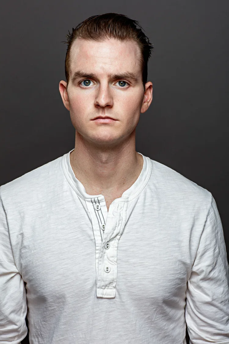 Portrait of a young man with fair skin, light brown hair, and blue eyes, wearing a white henley shirt, against a dark gray background.