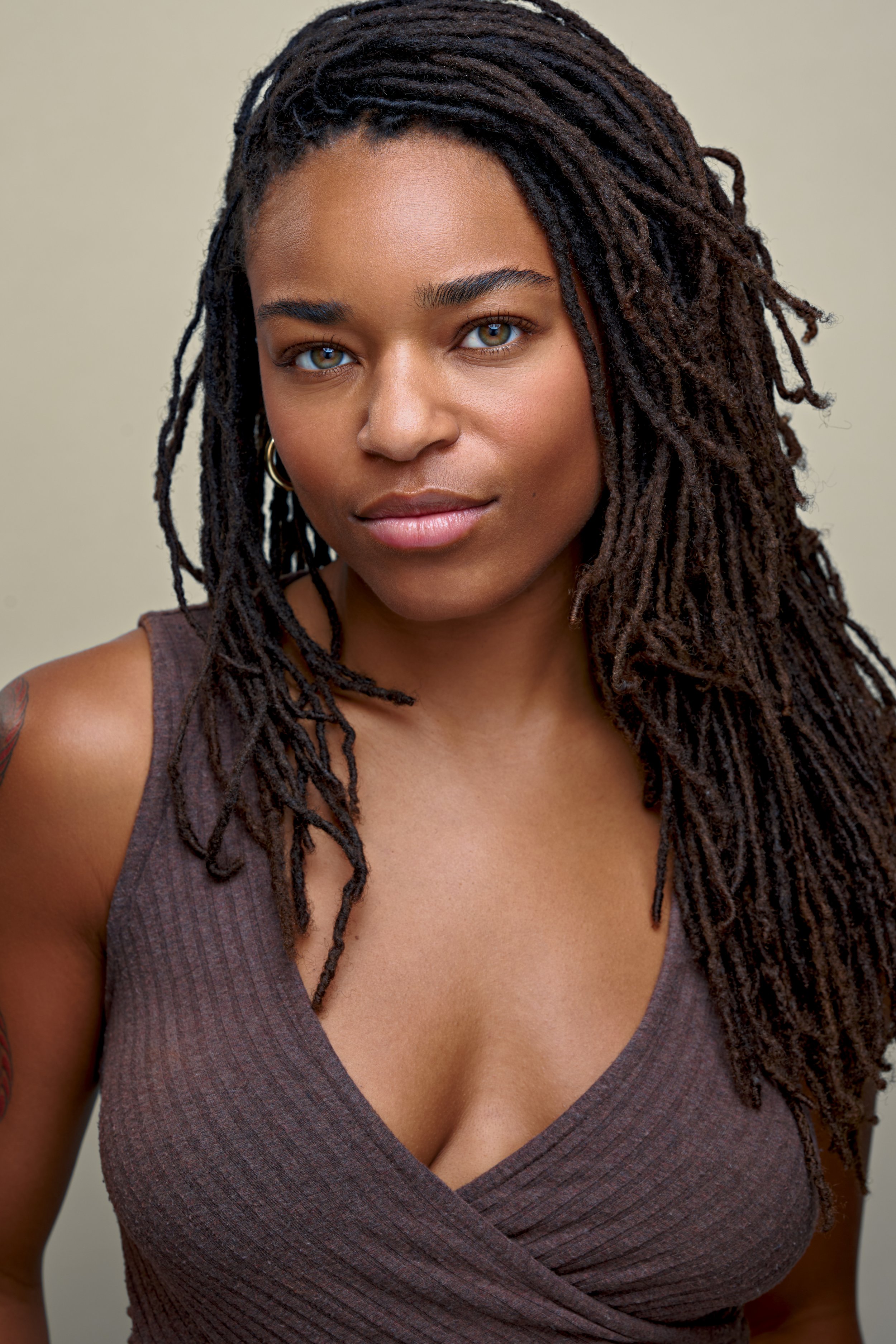 Close-up of a woman with long, textured dreadlocks, wearing a sleeveless brown top with a deep V-neck, looking directly at the camera with a neutral expression.