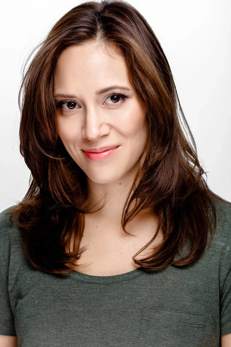 A woman with brown hair and fair skin wearing a dark green top, smiling in front of a plain white background.