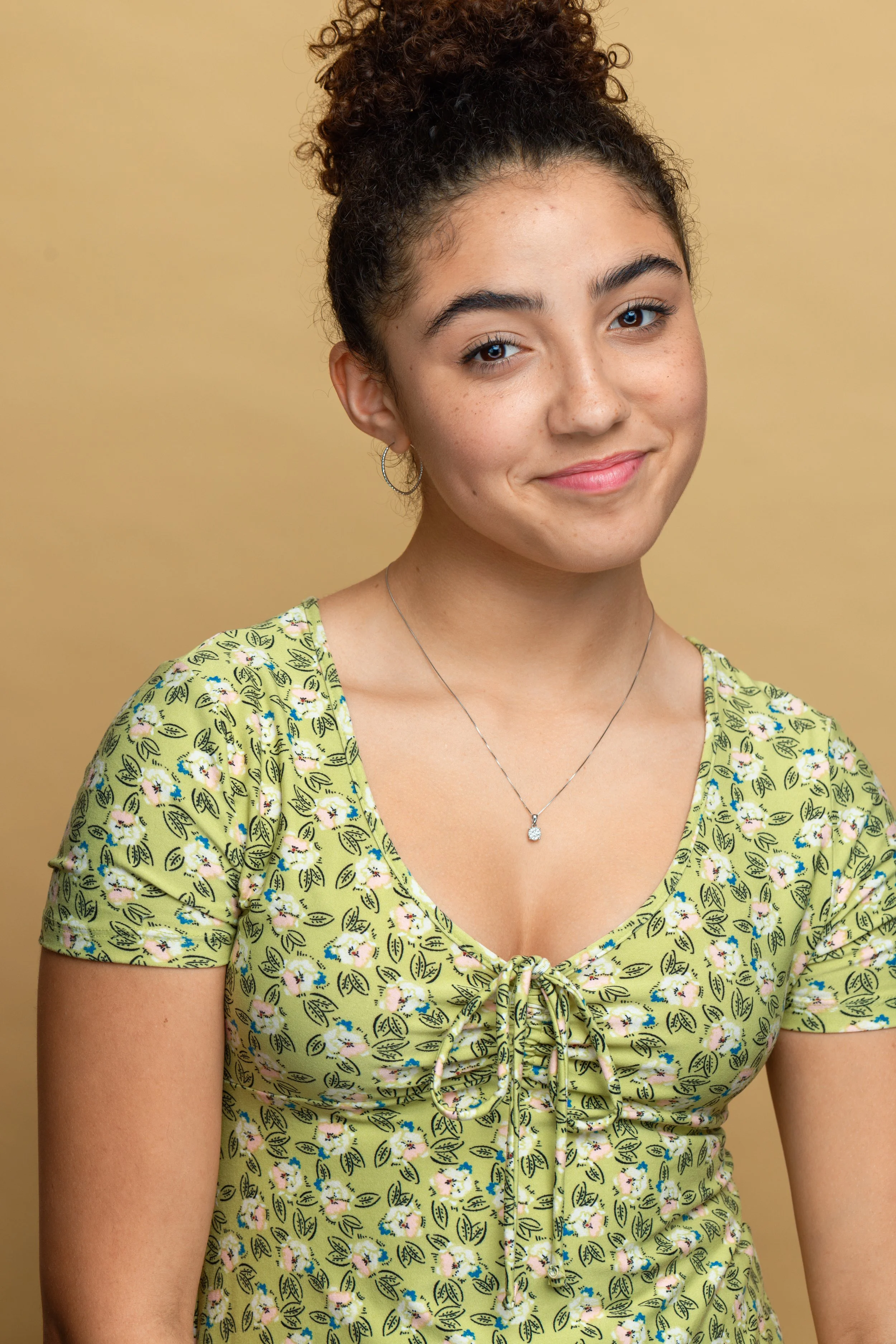 A young woman with curly brown hair tied up, wearing a yellow floral dress with a tie at the front, and silver jewelry, smiling against a plain yellow background.