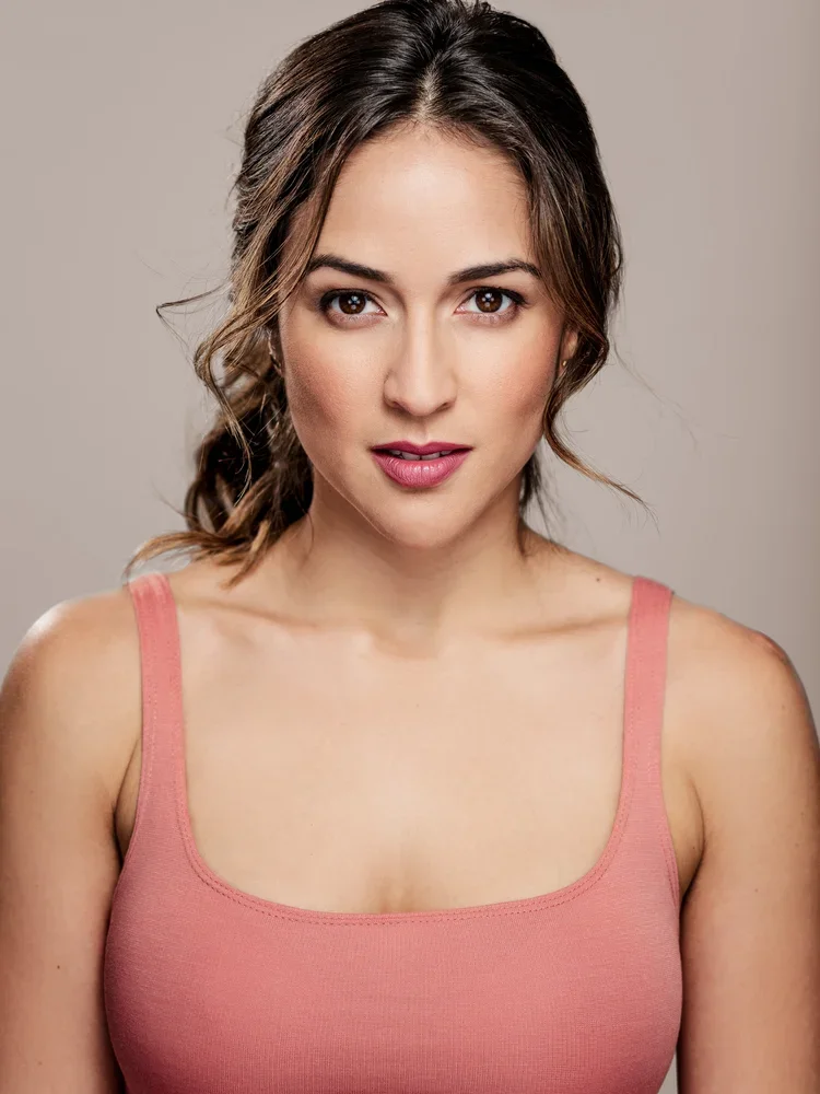 A young woman with brown hair styled in loose waves, wearing a pink tank top, looking directly at the camera with a soft expression against a neutral background.