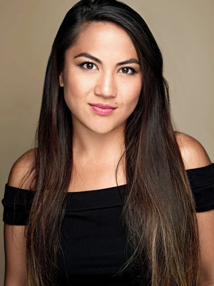 Portrait of a young woman with long brown hair, wearing a black off-the-shoulder top, smiling slightly against a neutral background.