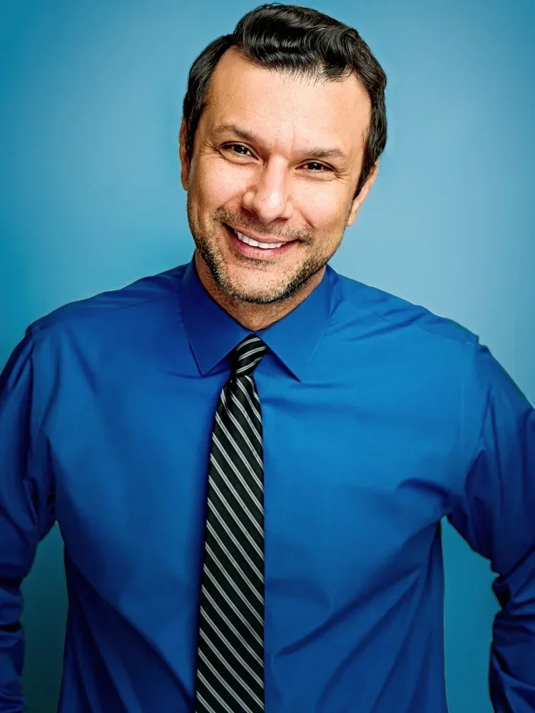 Smiling man with dark hair and a beard, wearing a blue dress shirt and a striped black and gray tie, standing against a blue background.