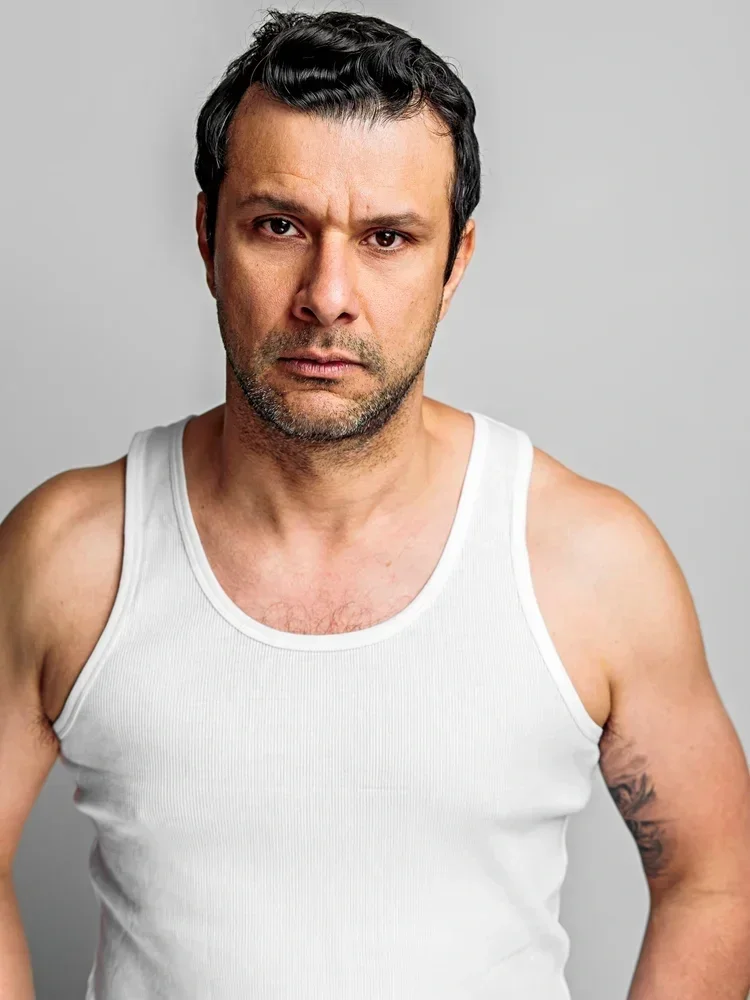 A man with dark hair and a serious expression, wearing a white tank top, standing against a plain gray background.