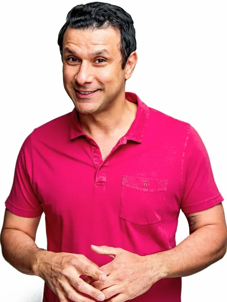 A man with dark hair and light skin wearing a bright pink short-sleeve collared shirt, smiling and standing with hands clasped in front of him against a white background.