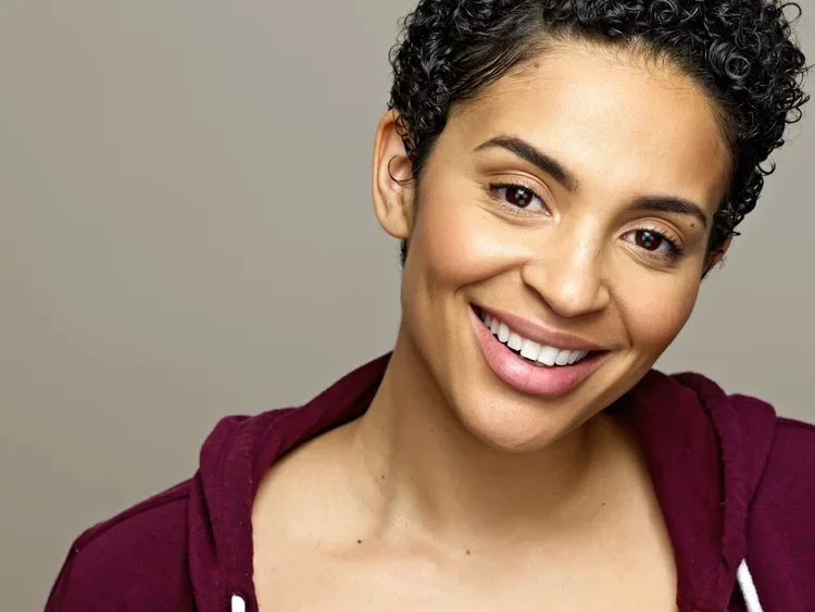 Close-up of a woman with short curly hair smiling at the camera wearing a maroon hoodie.