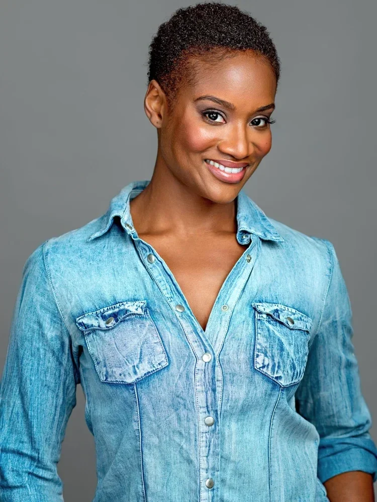 A smiling woman with short curly black hair wearing a light blue denim shirt against a plain gray background.