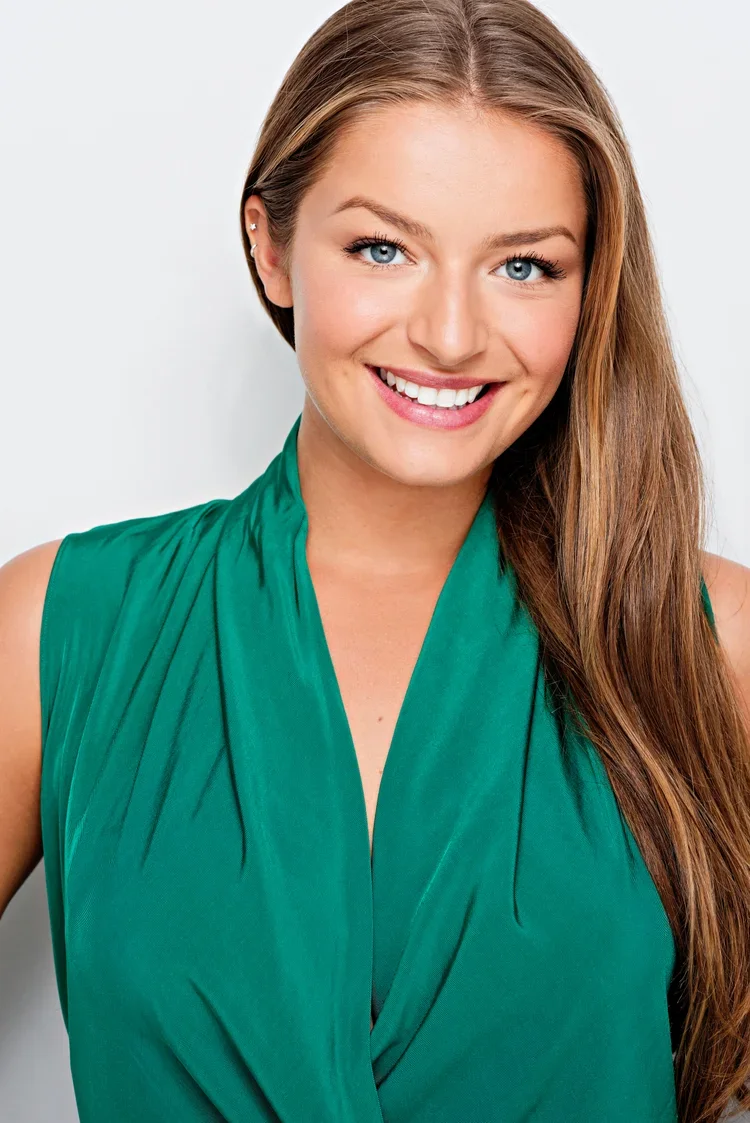 A smiling woman with long brown hair and blue eyes wearing a sleeveless green top against a plain white background.