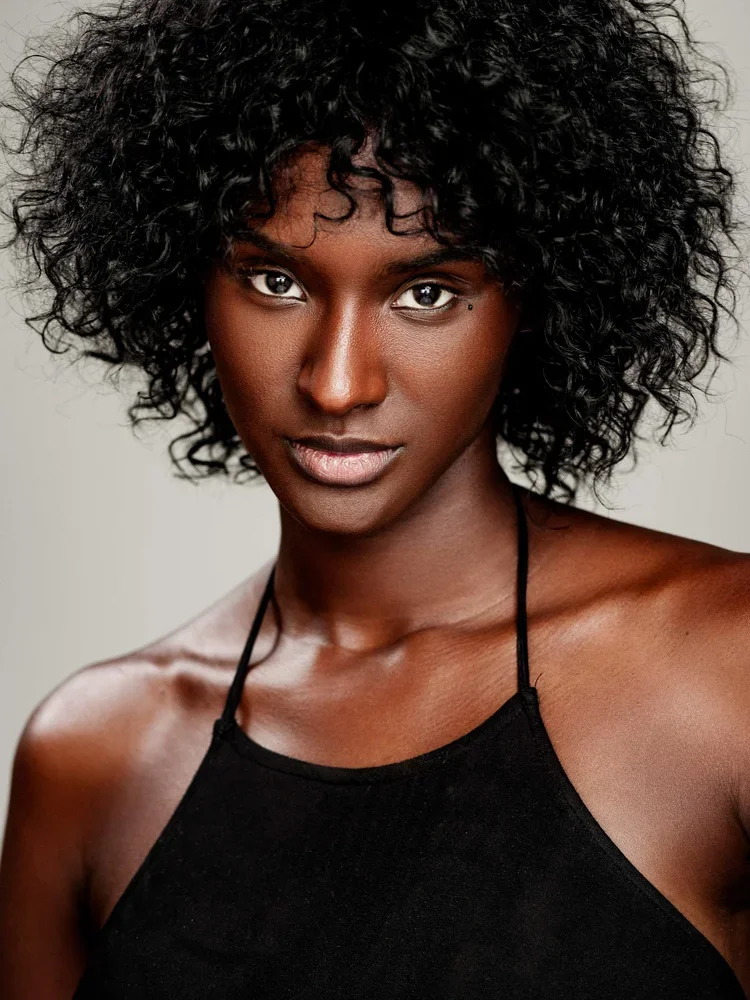 Close-up portrait of a Black woman with curly black hair, wearing a black tank top, looking directly at the camera against a neutral background.