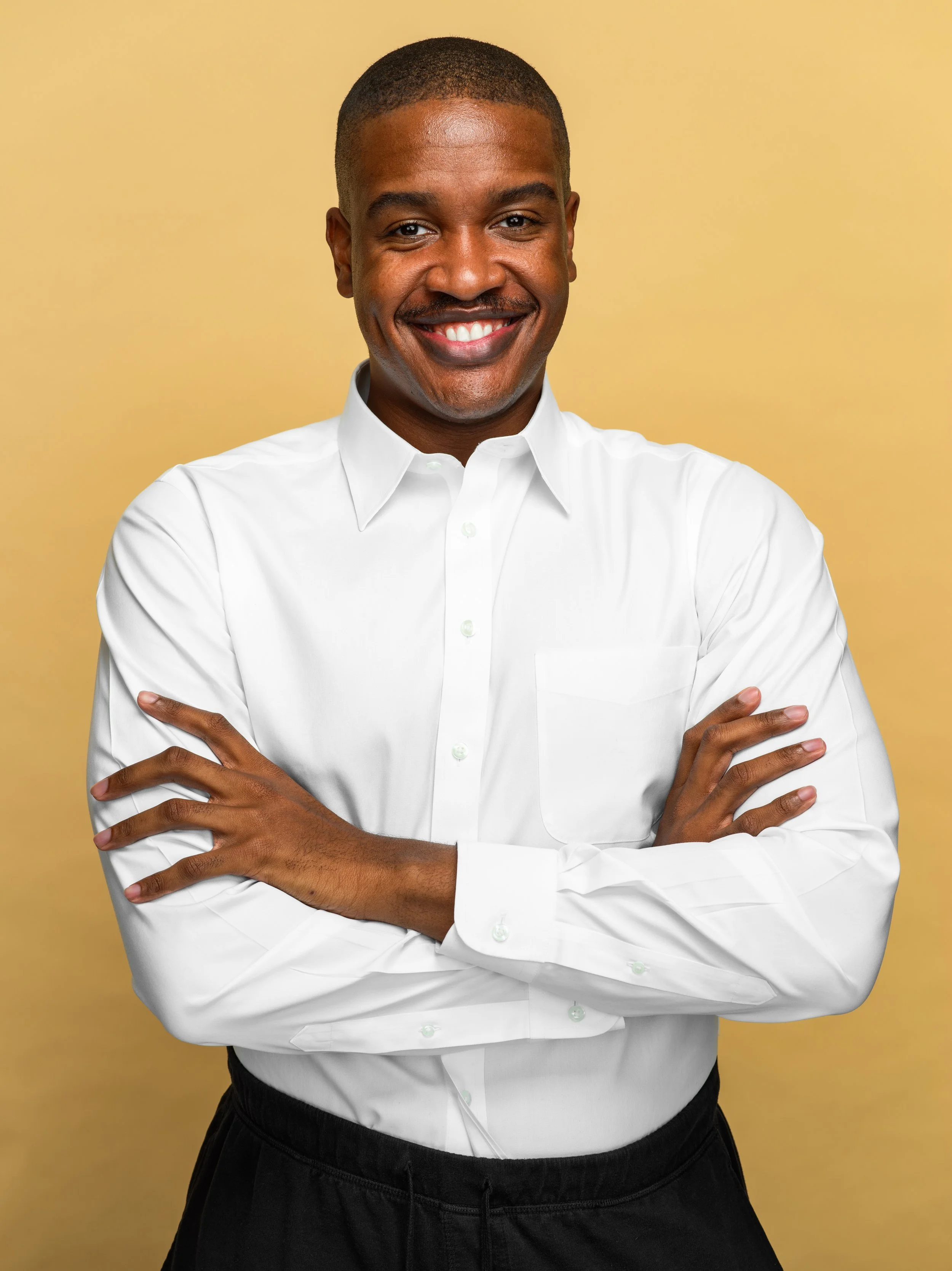 A smiling man with crossed arms, wearing a white dress shirt and black pants, standing against a yellow background.