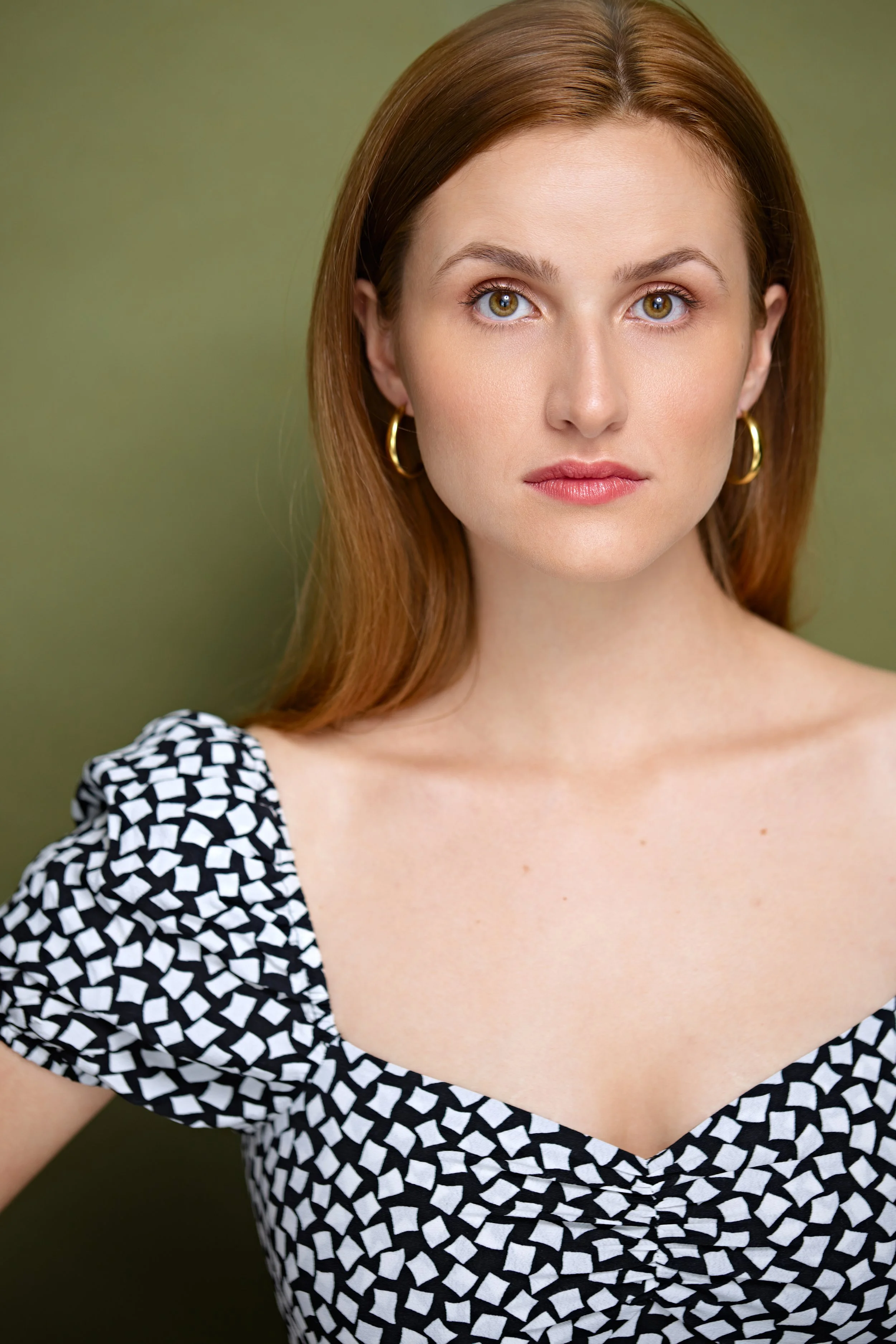 A woman with reddish-brown hair, wearing gold hoop earrings and a black and white patterned dress with puffed sleeves, looking directly at the camera with a neutral expression, against a plain green background.