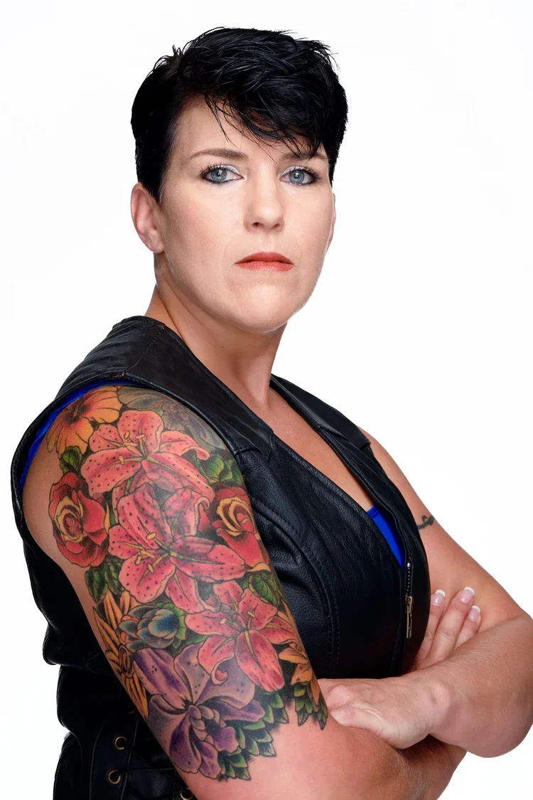 A woman with short black hair, blue eyes, and a serious expression, wearing a black leather vest and a blue top, with a colorful floral tattoo sleeve on her left arm against a plain white background.