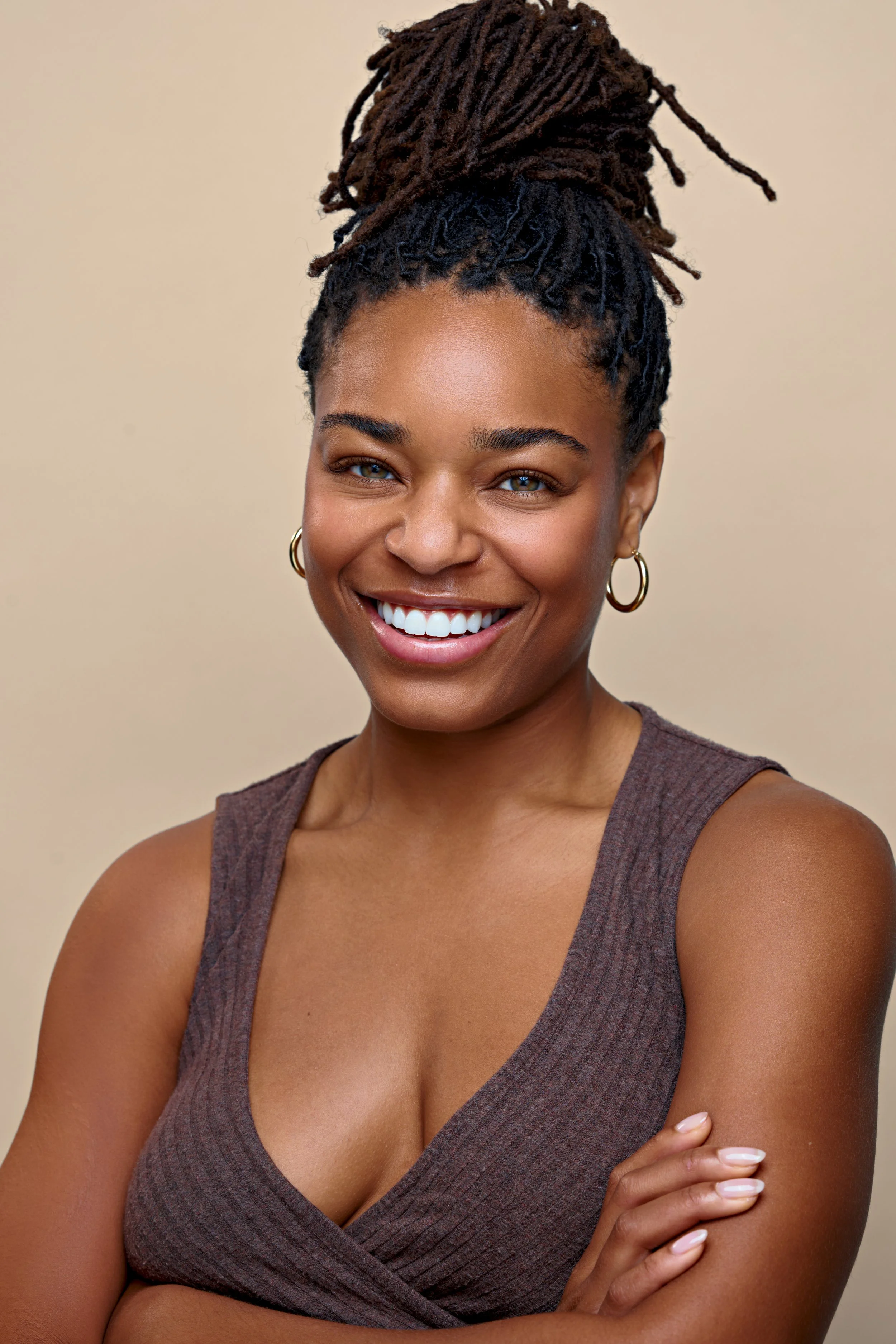 A woman with a dark complexion and brown dreadlocks styled in a high bun, smiling with white teeth, wearing gold hoop earrings, and a sleeveless brown top, standing against a plain beige background.