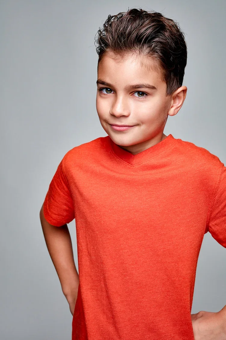 A young boy with short dark hair, blue eyes, and a slight smile, wearing an orange t-shirt against a plain gray background.