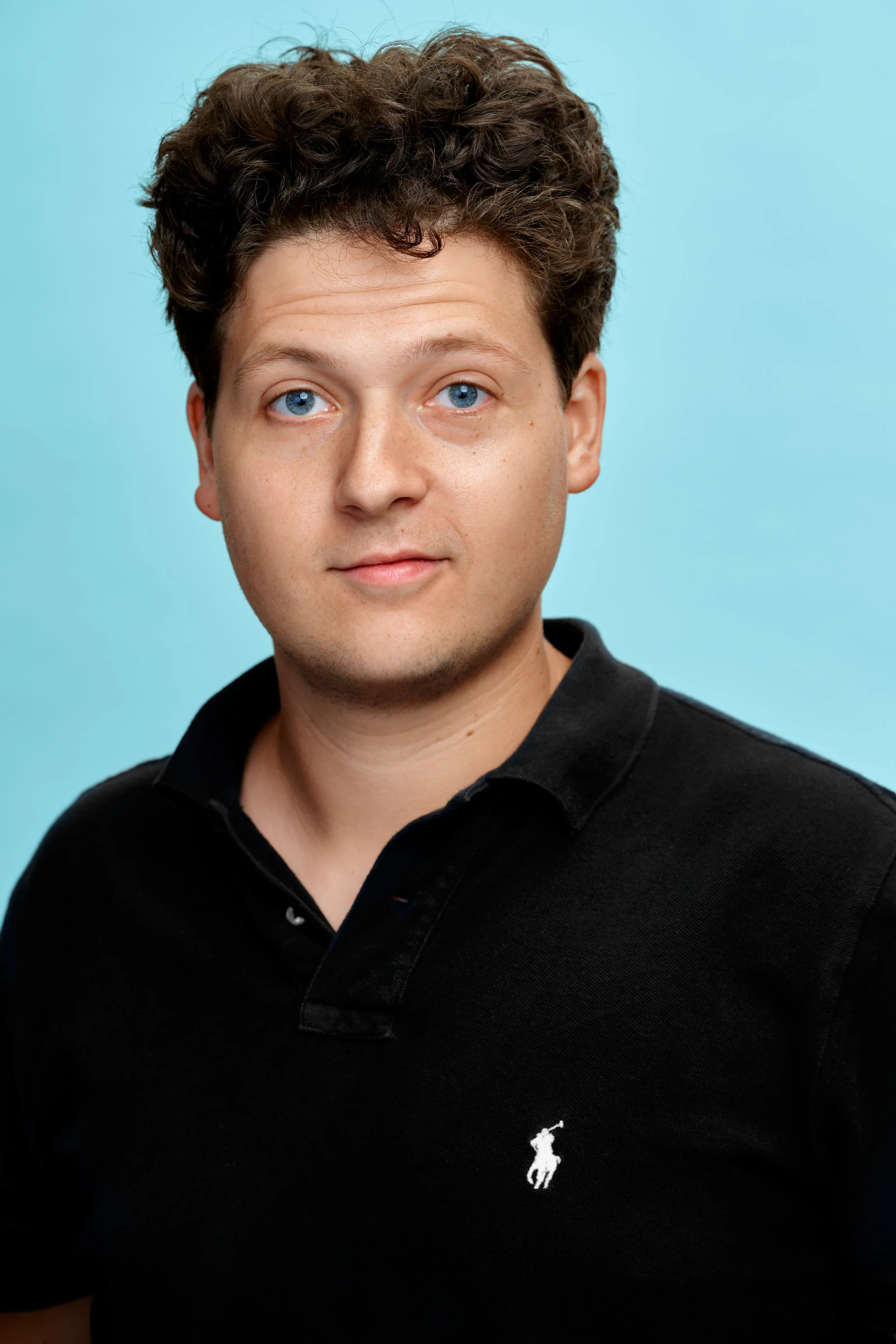 A man with curly brown hair and blue eyes wearing a black polo shirt with a white embroidered logo of a horse and rider, against a light blue background.