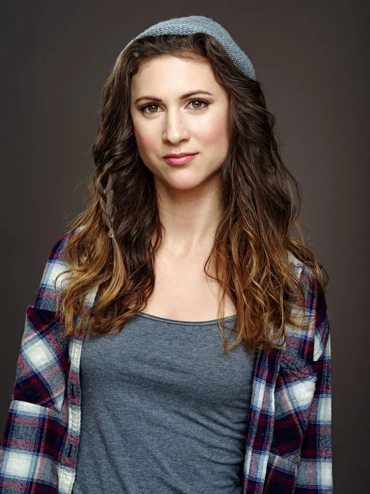 A young woman with long, wavy brown hair, wearing a gray beanie, gray shirt, and a plaid shirt, against a dark background.