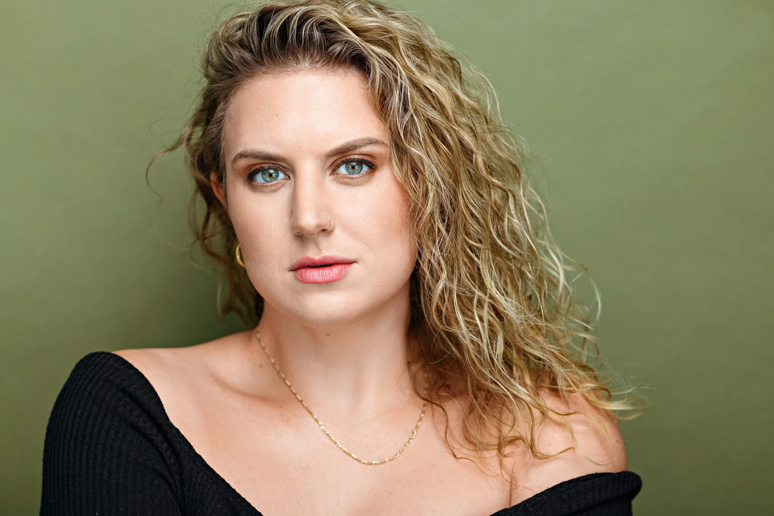Close-up portrait of a woman with curly blonde hair, blue eyes, and a nose ring, wearing a black off-the-shoulder top, gold jewelry, and a subtle smile, against a green background.