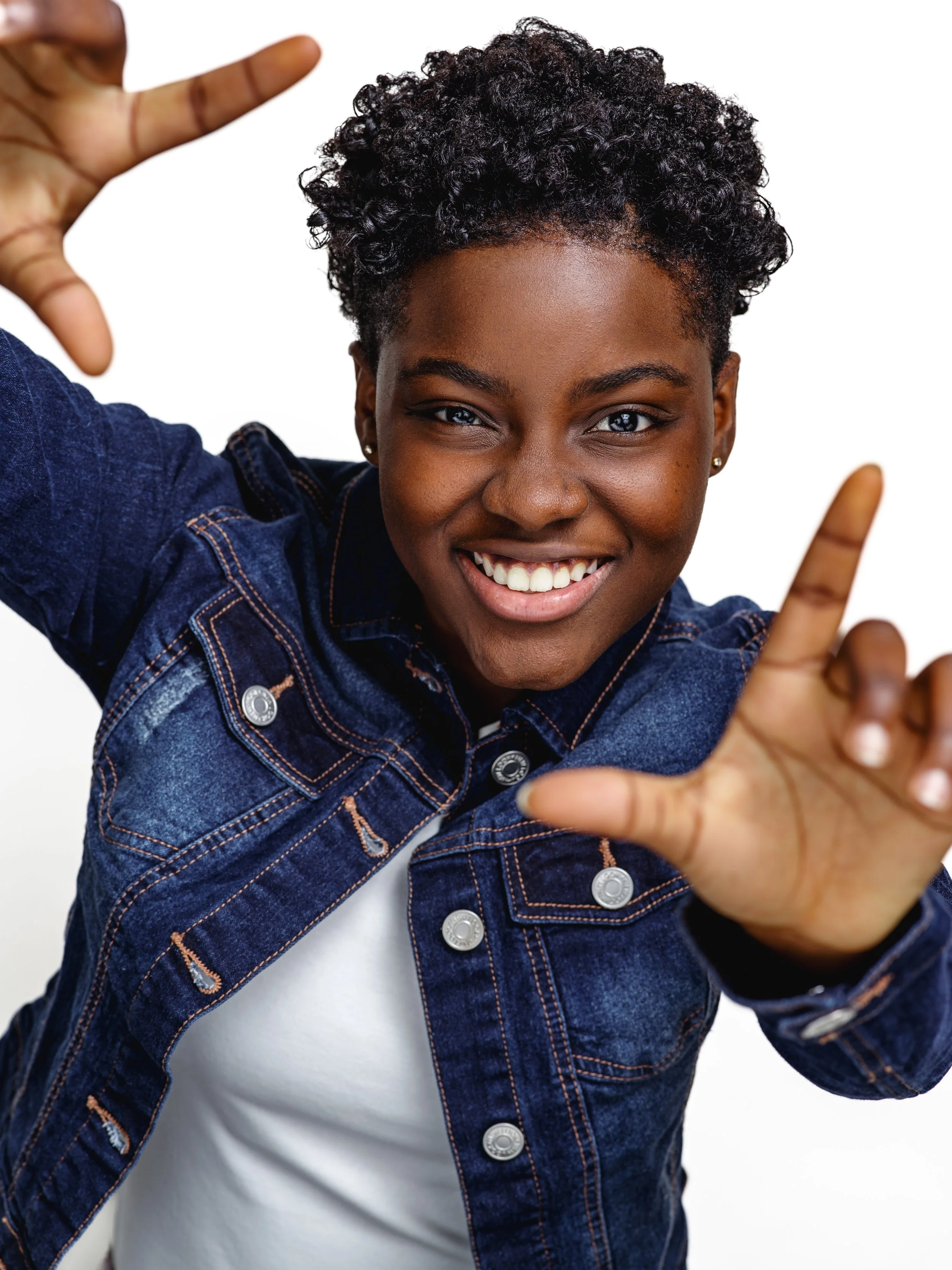 A young woman with short curly hair smiling at the camera while making a framing gesture with her hands.