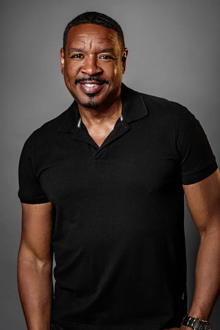 Portrait of a smiling middle-aged African American man with short black hair, wearing a black polo shirt, against a gray background.