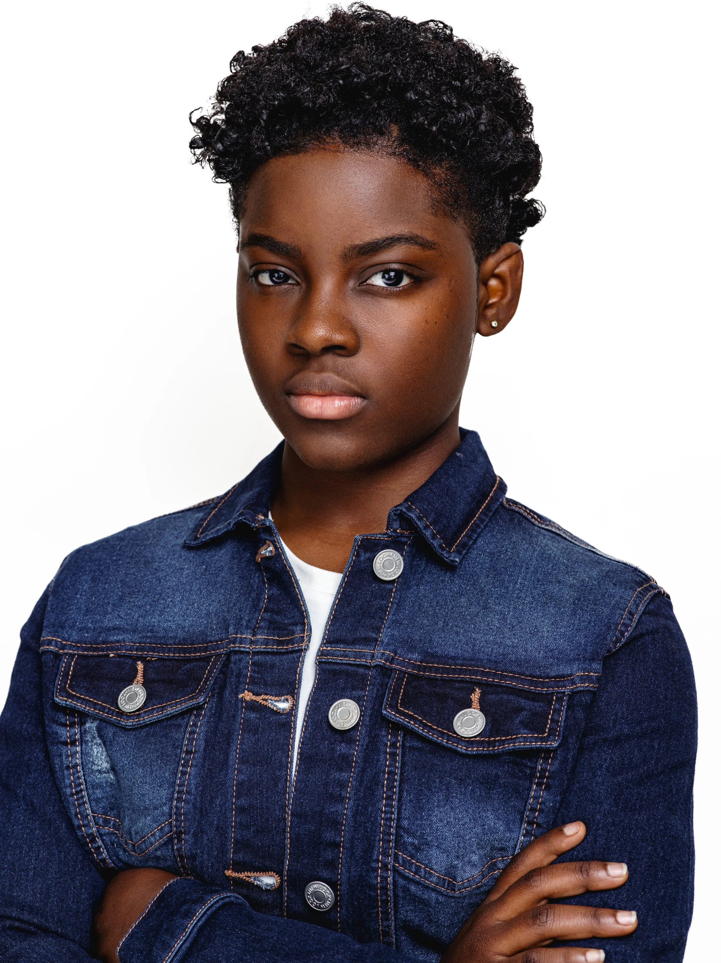 A young Black woman with short, curly hair wearing a dark denim jacket and white shirt, standing against a white background.
